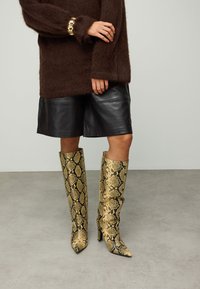 Goudkleurige en zwarte slangenprint kniehoge laarzen met een puntige neus en naaldhak, gecombineerd met zwarte leren shorts en een bruine, pluizige trui.