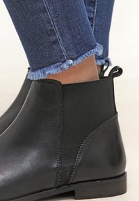 Black ankle boots made of smooth leather with elastic side panels, flat heel, and contrast stitching, paired with frayed hem denim jeans.