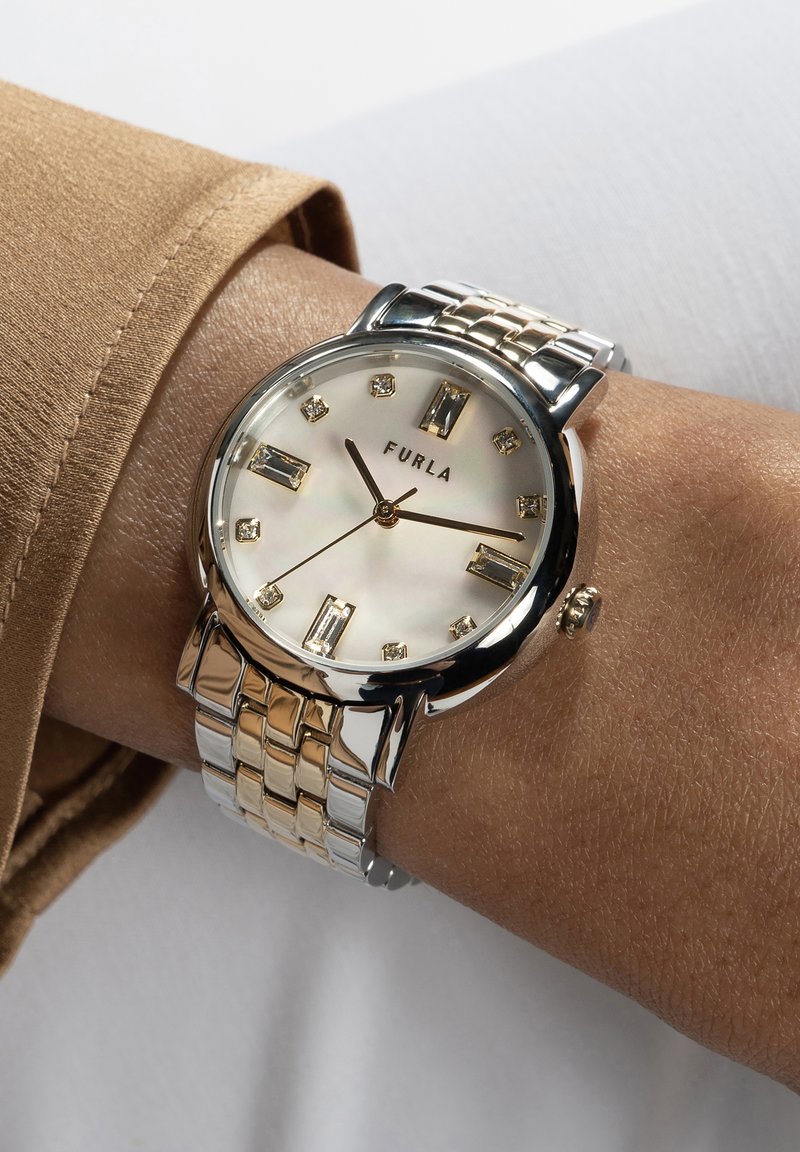 Reloj Furla con una correa metálica redonda de plata y oro, cara de madreperla blanca y marcadores de hora rectangulares. Cuenta con una corona de presión.
