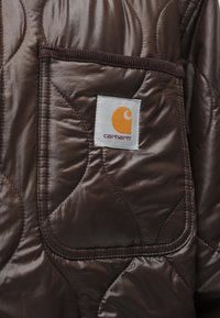Hnědá prošívaná bunda s výraznou kapsou a nášivkou loga Carhartt v oranžové a bílé barvě, s hladkým a lesklým povrchem.