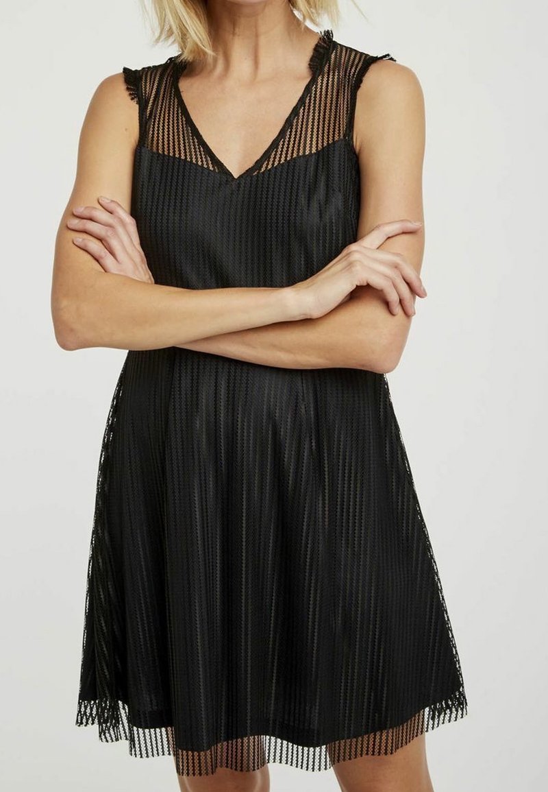 Femme portant une robe noire en résille sans manches, bras croisés, sur un fond clair uni.