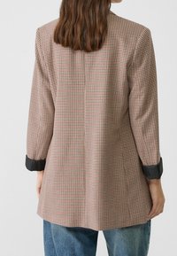 Blazer a cuadros en marrón, crema y negro. Presenta mangas largas con puños negros y una silueta entallada. La tela parece texturada.