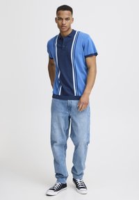 Kék póló sötét tengeri kék és világos kék csíkokkal, rövid ujjú és galléros. Világos kék farmernadrággal és fekete sportcipőkkel viselve.