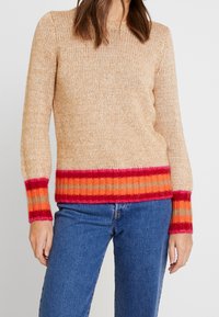 Stickad tröja i ljus beige med en flerfärgad randig fåll och ärmar i rött, orange och rosa. Strukturerad tyg med långa ärmar.