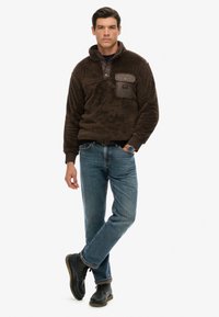 Brun fleecepullover med halvlukket front, brystlomme i mørkt stof, parret med blå jeans og sorte snørebåndsstøvler.