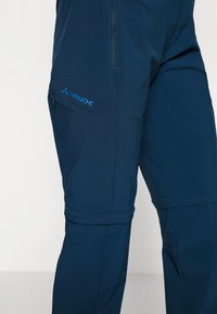 Marineblauwe broek met een ritszak aan de zijkant, met een gladde textuur en een logodetail. De broek heeft een taps toelopend ontwerp, geschikt voor gebruik buitenshuis.