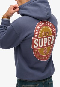 Man in een blauwe hoodie met een groot rond logo in rood en mosterdkleur op de achterkant, met de tekst "Premium Quality Superdry."