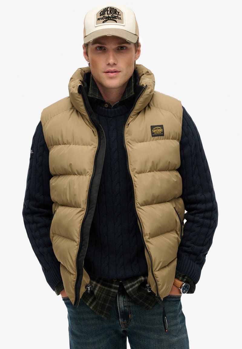 Veste sans manches matelassée couleur fauve avec un col montant, fermeture éclair et écusson logo, portée sur un pull en maille torsadée bleu marine et une chemise à carreaux verte.
