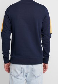 Felpa blu navy con strisce gialle e bianche a contrasto sulle maniche, colletto e polsini a coste; texture morbida e vestibilità casual.