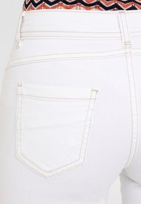 Weiße Denim-Jeans haben eine sichtbare Gesäßtasche mit einem dezenten Stichdetail. Der Stoff wirkt glatt mit einer leichten Textur.