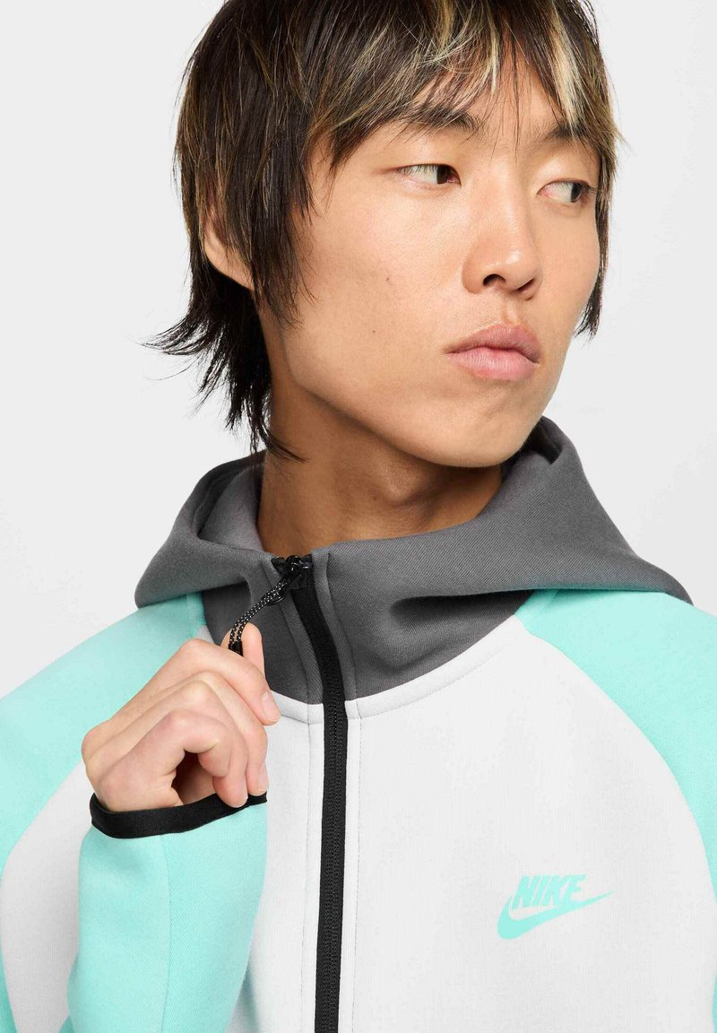 Chaqueta con capucha y cremallera en gris, blanco y menta. Presenta una textura suave, colores contrastantes y un pequeño logo de Nike en el pecho.