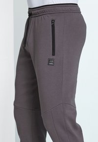 Gray jogger pants made of soft fabric, featuring a zippered side pocket, elastic waistband, and logo patch on the thigh.