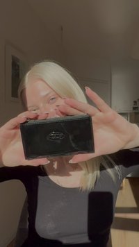 Ung kvinna med blont hår som håller en svart plånbok i lackläder med silverlåsanordning, inomhus i naturligt solljus.