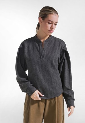 Kvinde med håret bundet tilbage iført mørkegrå bluse med lange ærmer og v-hals samt brune bukser, ser ned og justerer sin bluse.