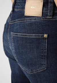 Donkerblauwe spijkerbroek met een gouden knoopaccent, contrasterende stiksels en een tan leren logo-opname bij de tailleband.