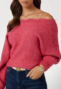 Pull en maille fuchsia à épaules dénudées avec une finition texturée et une coupe ample, associé à un jean noir et à un bracelet en chaîne dorée.
