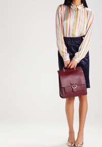 Sac à dos en cuir bordeaux avec un rabat et des accessoires ronds, associé à un chemisier à rayures pastel et une jupe en satin bleu marine.