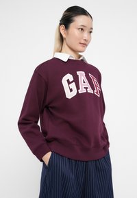 Vrouw met licht haar in een lage bun, gekleed in een bordeauxrood GAP-sweater over een witte blouse en marineblauwe gestreepte broek, met handen in de zakken.