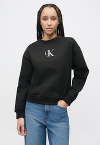 Czarna bluza z okrągłym dekoltem, z małym szarym logo "cK" na piersi. W zestawie z jasnoniebieskimi jeansami.