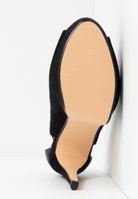 Chaussure à talon haut en daim noir, inclinée vers le haut, montrant une semelle beige lisse et un talon étroit sur une surface et un fond blancs.