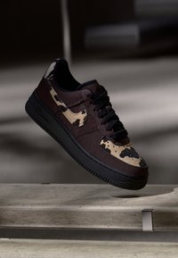 Zapatillas de deporte marrón oscuro con un acabado texturizado, que presentan una suela negra y un patrón beige con manchas en los laterales. Cuerdas negras y marca visibles.