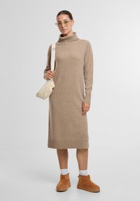 Beiges Strickkleid mit hohem Kragen, gerippten Bündchen und Saum. Kombiniert mit braunen Knöchelboots und einer gesteppten weißen Umhängetasche.