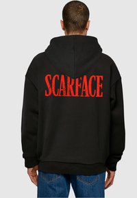 Crna kapuljača s velikim crvenim grafičkim natpisom "SCARFACE" na stražnjem dijelu, mekog materijala i opuštenog kroja.