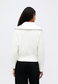 Biely rebrovaný pletený sveter s nadrozmerným sklápacím golierom, priliehavým pásom a dlhými rukávmi; doplnený o čierne nohavice s vysokým pásom.