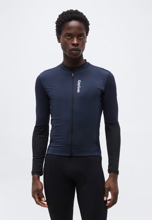 Νέος άνδρας που φοράει μπλε σκούρη και μαύρη μακρυμάνικη ποδηλατική μπλούζα με φερμουάρ μπροστά και το λογότυπο GripGrab στο στήθος, στέκεται μπροστά σε μονόχρωμο φόντο.