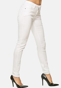 Jeans blancos ajustados hechos de una mezcla de algodón elástico. Presentan una cremallera en la parte delantera y un diseño elegante, combinados con tacones altos de charol negros.