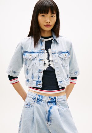 Joven mujer con chaqueta vaquera corta azul claro y vaqueros de talle alto a juego, sobre una camiseta oscura con estampado y puños a rayas.