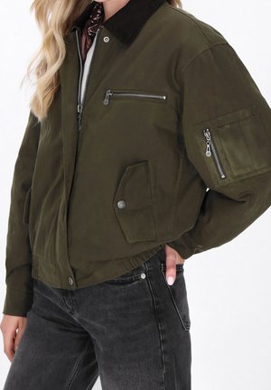 Personne portant un blouson bomber vert olive avec des fermetures éclair et un jean foncé, main gauche dans la poche, longs cheveux blonds ondulés partiellement visibles.