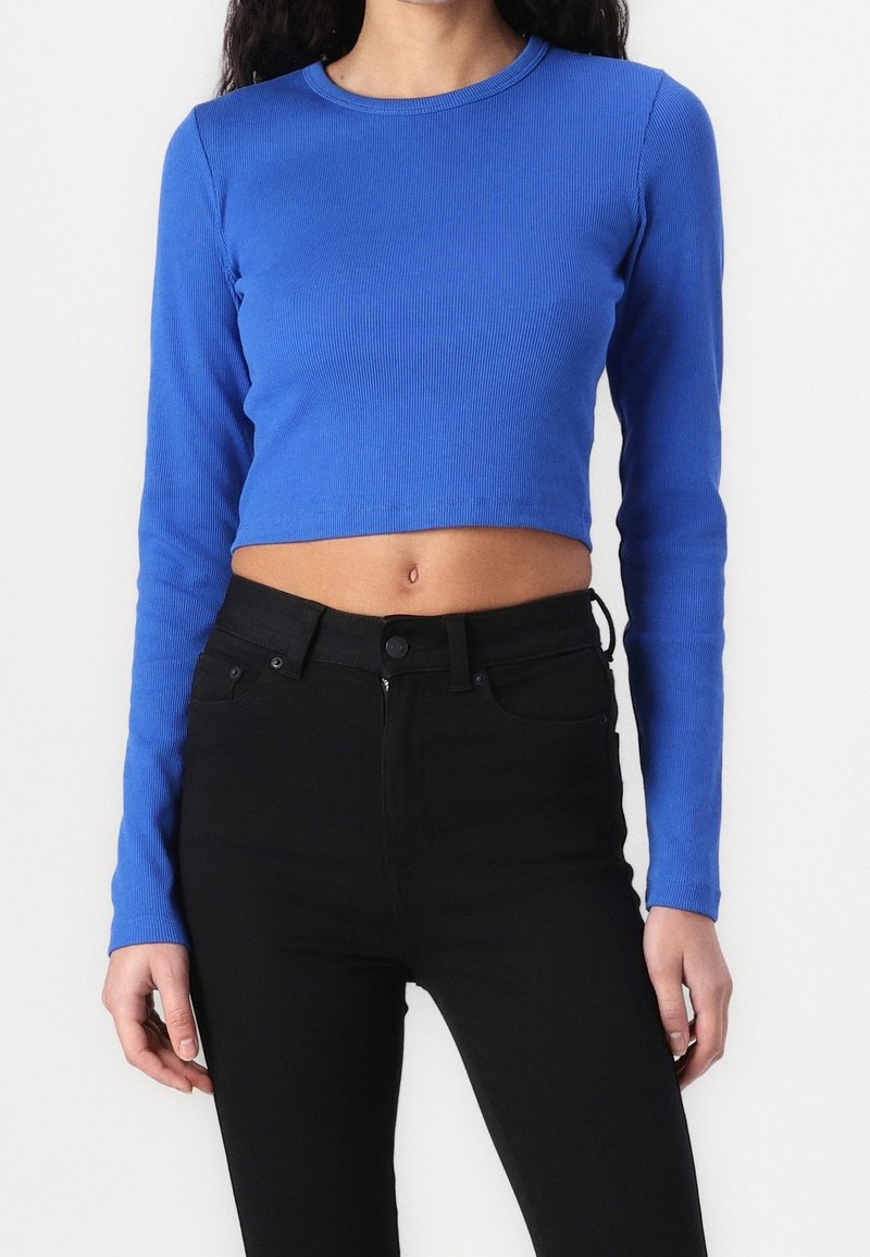 Long-sleeve cropped top in royal blue ribbed fabric. Features a round neckline and fitted sleeves, paired with black high-waisted jeans.