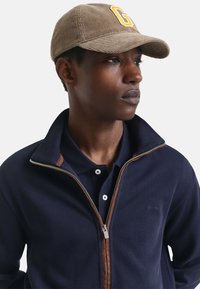 Gorra de pana marrón con emblema amarillo "G", chaqueta azul marino con detalles en cremallera marrón y camiseta de polo azul marino con dos botones.