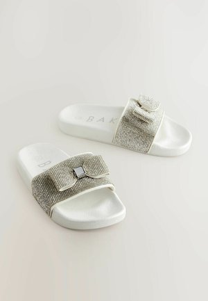 Baltās slaidas sandales ar teksturētu siksniņu, kas pārklāta ar mirdzošām rhinestone akmentiņiem, un dekoratīvu loku ar kvadrātveida akcentu.