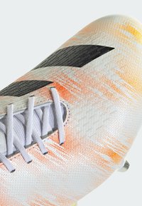Botín de fútbol con una parte superior texturizada y multicolor en blanco, naranja y amarillo. Presenta cordones plateados y un acento de logo negro.