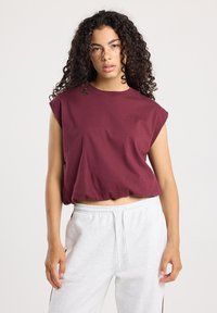 Jeune femme aux cheveux bouclés, portant un haut sans manches bordeaux et un pantalon de survêtement gris clair, se tenant devant un fond uni.