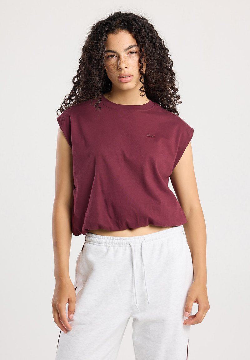 Jeune femme aux cheveux bouclés, portant un haut sans manches bordeaux et un pantalon de survêtement gris clair, se tenant devant un fond uni.