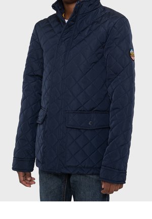 Granatowa pikowana kurtka z wysokim kołnierzem, zapięciem na zatrzaski i przednimi kieszeniami. Na górnym rękawie znajduje się naszywka. Gładka tekstura.