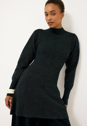 Donna che indossa un abito a maglia a costine aderente di colore grigio scuro con maniche lunghe e collo alto, posando contro un muro bianco.
