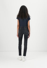 Jeans ajustados negros de talle alto con un desvanecimiento sutil, combinados con una blusa corta de manga corta en color azul marino; la modelo está de pie con zapatillas blancas, mostrando la vista desde atrás.