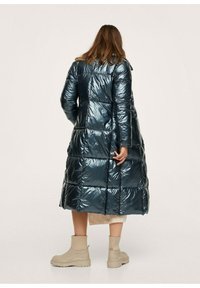 Lang dynejakke i mørk teal, lavet af blankt materiale med et quiltet design. Har en knælængde og sidelommer. Et par beige støvler er synlige.