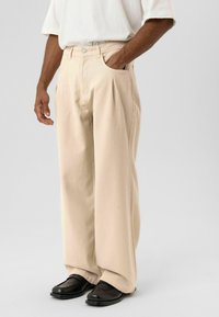 Beige wijd uitlopende broek van katoen, met plooien aan de voorkant en zijzakken. Gecombineerd met zwarte loafers voor een casual uitstraling.