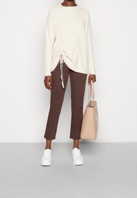 Maglione oversized color beige chiaro con arricciature laterali, pantaloni cropped marroni, sneakers bianche e una borsa color beige chiaro. Texture liscia.