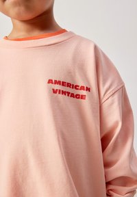 Enfant portant un sweat-shirt rose clair avec un texte rouge "AMERICAN VINTAGE" sur la poitrine et un col orange en dessous.