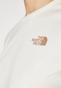 Kermansävyinen puuvillapaita, jossa on vaaleanruskea "The North Face" -logo rinnan vasemmalla puolella. Lyhyet hihat ja pyöreä kaula-aukko.