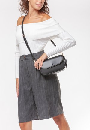 Borsa a tracolla nera in pelle con finitura liscia, caratterizzata da un design curvo e una tracolla sottile. Indossata da un modello, abbinata a pantaloni corti grigi a righe.