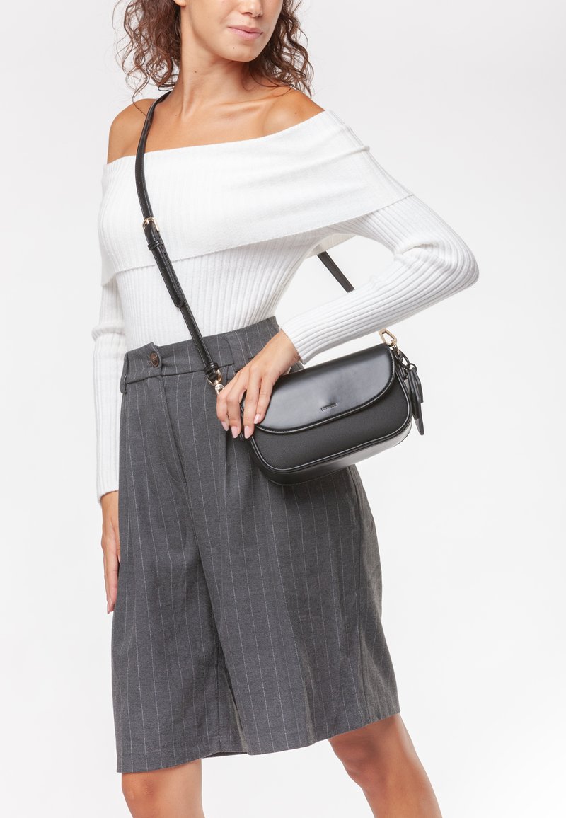 Borsa a tracolla nera in pelle con finitura liscia, caratterizzata da un design curvo e una tracolla sottile. Indossata da un modello, abbinata a pantaloni corti grigi a righe.
