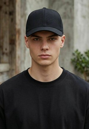 Young man with neutral expression wearing a black baseball cap and black shirt, standing against a blurred outdoor background.