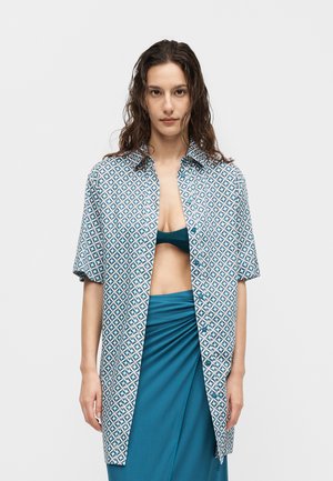 Kobieta o falowanych włosach, ubrana w niebieską, wzorzystą, oversize'ową koszulę otwartą na niebieskim topie bandeau i dopasowanej spódnicy z wysokim stanem na jednolitym tle.